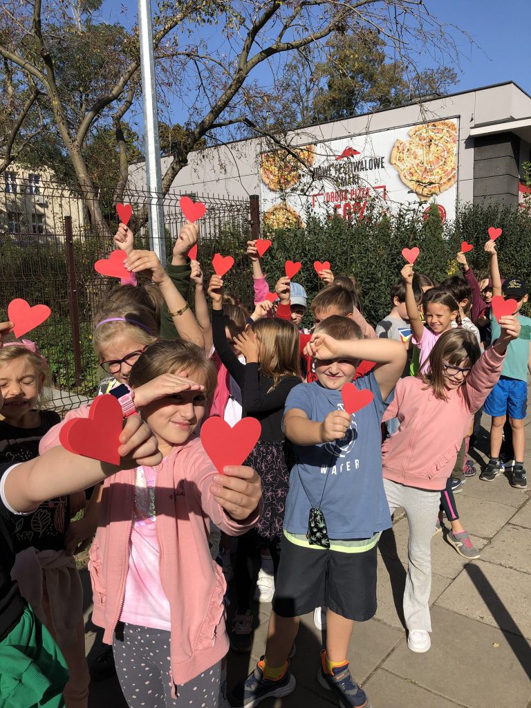 Duża grupa dzieci na szkolnym podwórku macha wyciętymi z papieru symbolami serca
