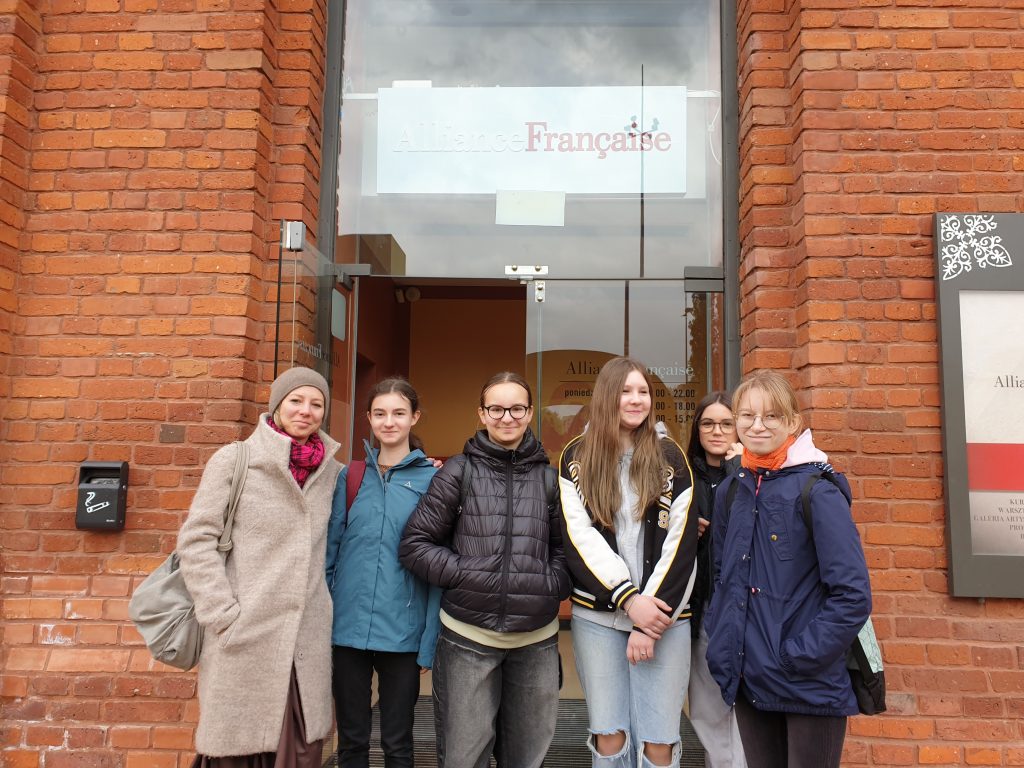 Grupa dziewcząt i nauczycielka pozują do zdjęcia przed wejściem do Alliance Française w Łodzi.