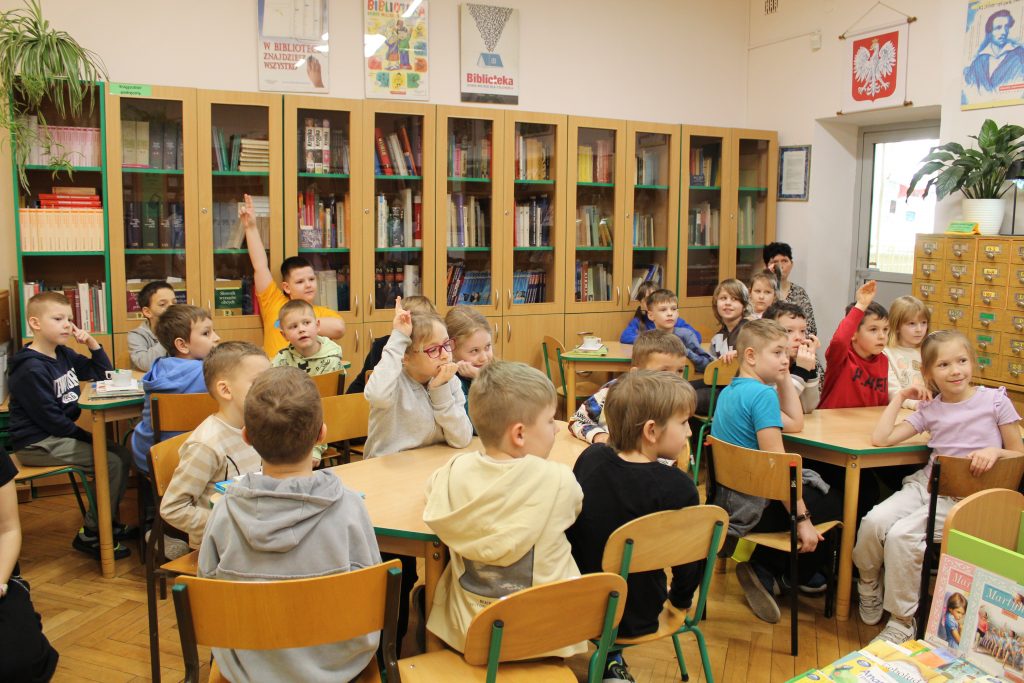 Grupa uczniów klas pierwszych siedząca przy stolikach. Niektóre dzieci podnoszą rękę.